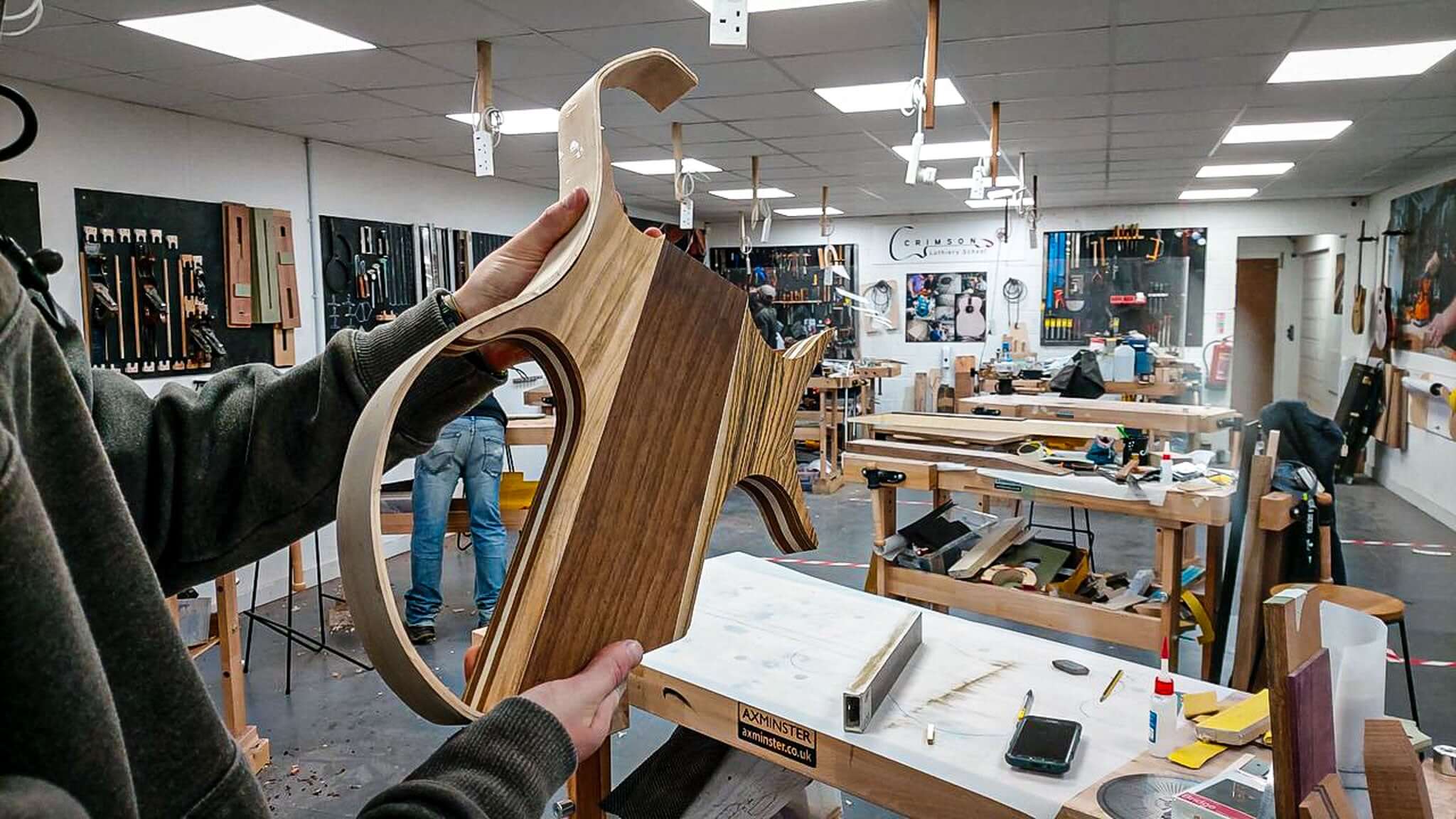 Craftsman holding a wooden guitar body in a busy workshop setting, showcasing handmade craftsmanship and woodworking skills.