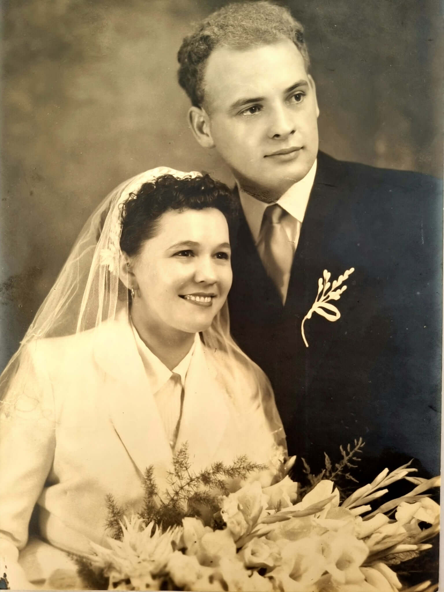 Vintage wedding photo of a bride in a veil and groom in a suit, holding a bouquet, with a sepia-toned finish.