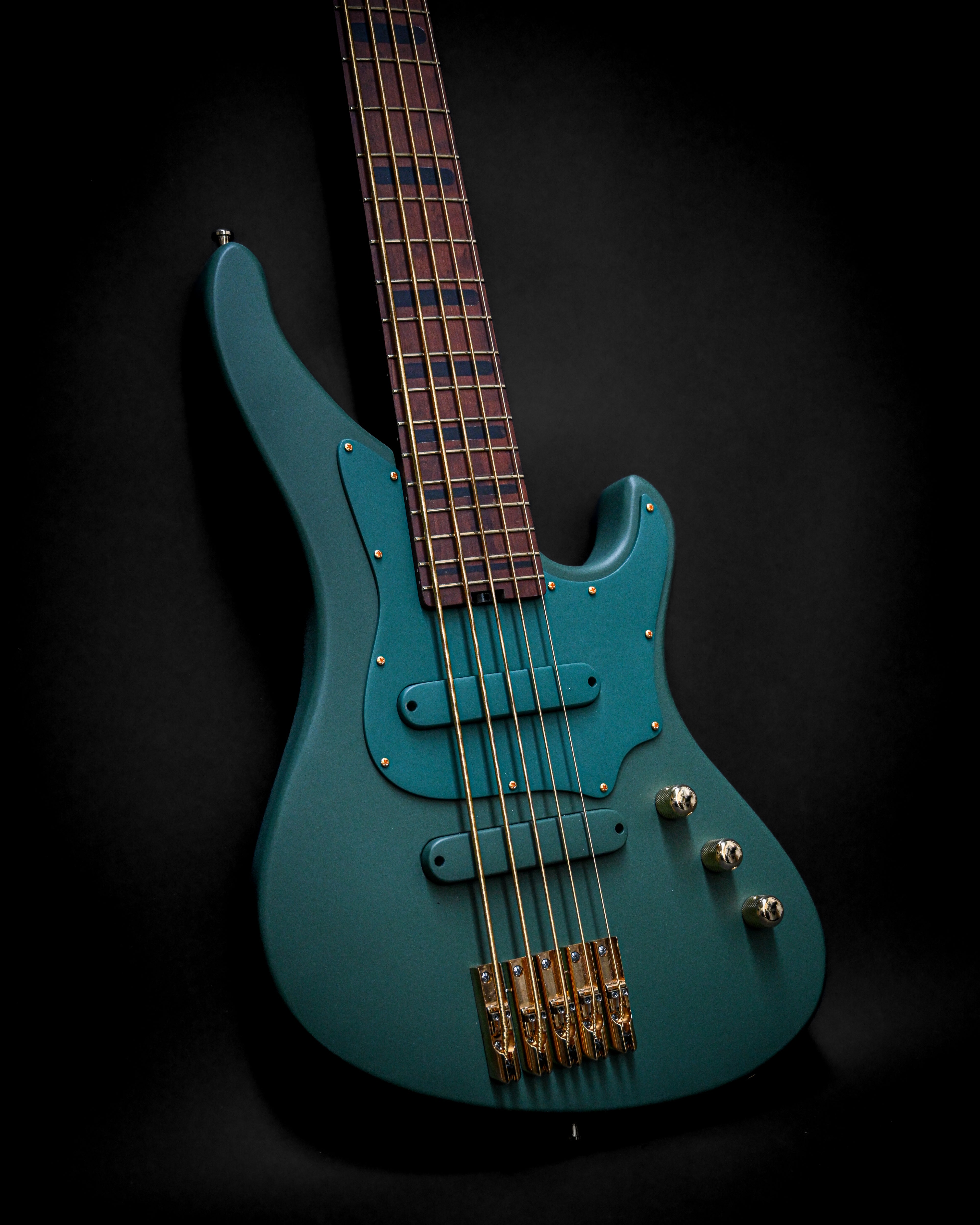 Electric blue bass guitar with five strings and gold tuning pegs against a dark background.
