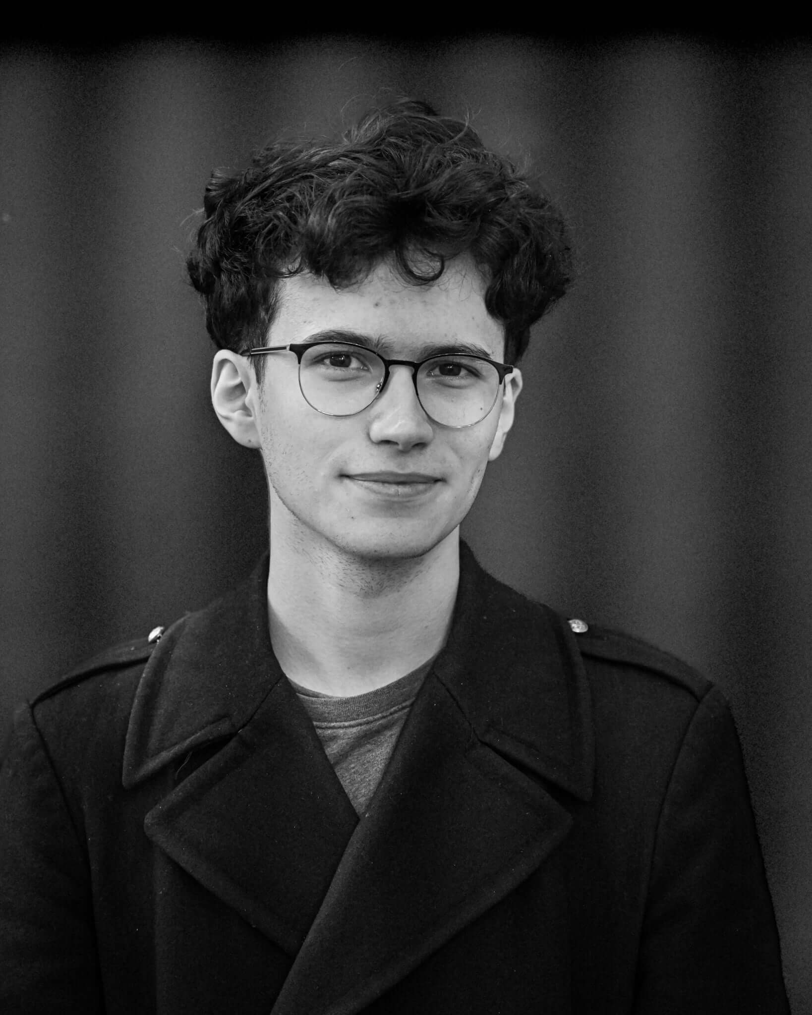 Young person with glasses and curly hair, wearing a dark coat, standing against a blurred background in black and white.