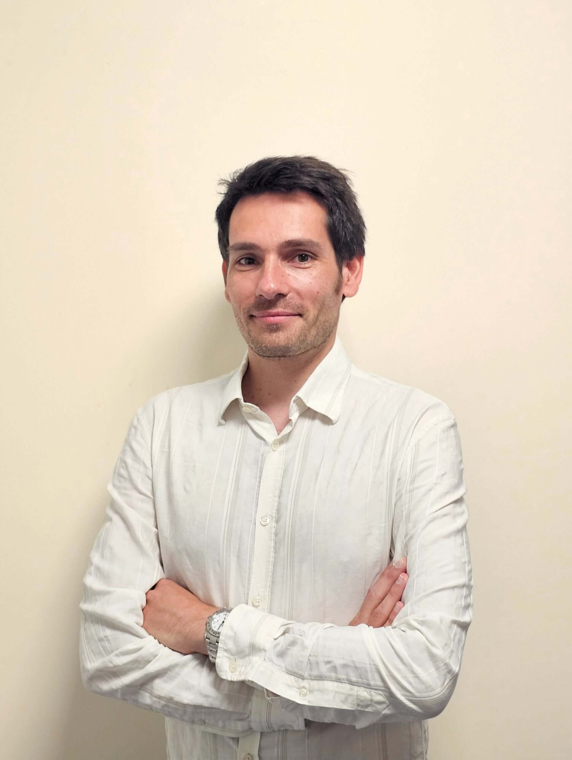 Persona con una camisa blanca de pie contra un fondo liso con los brazos cruzados.