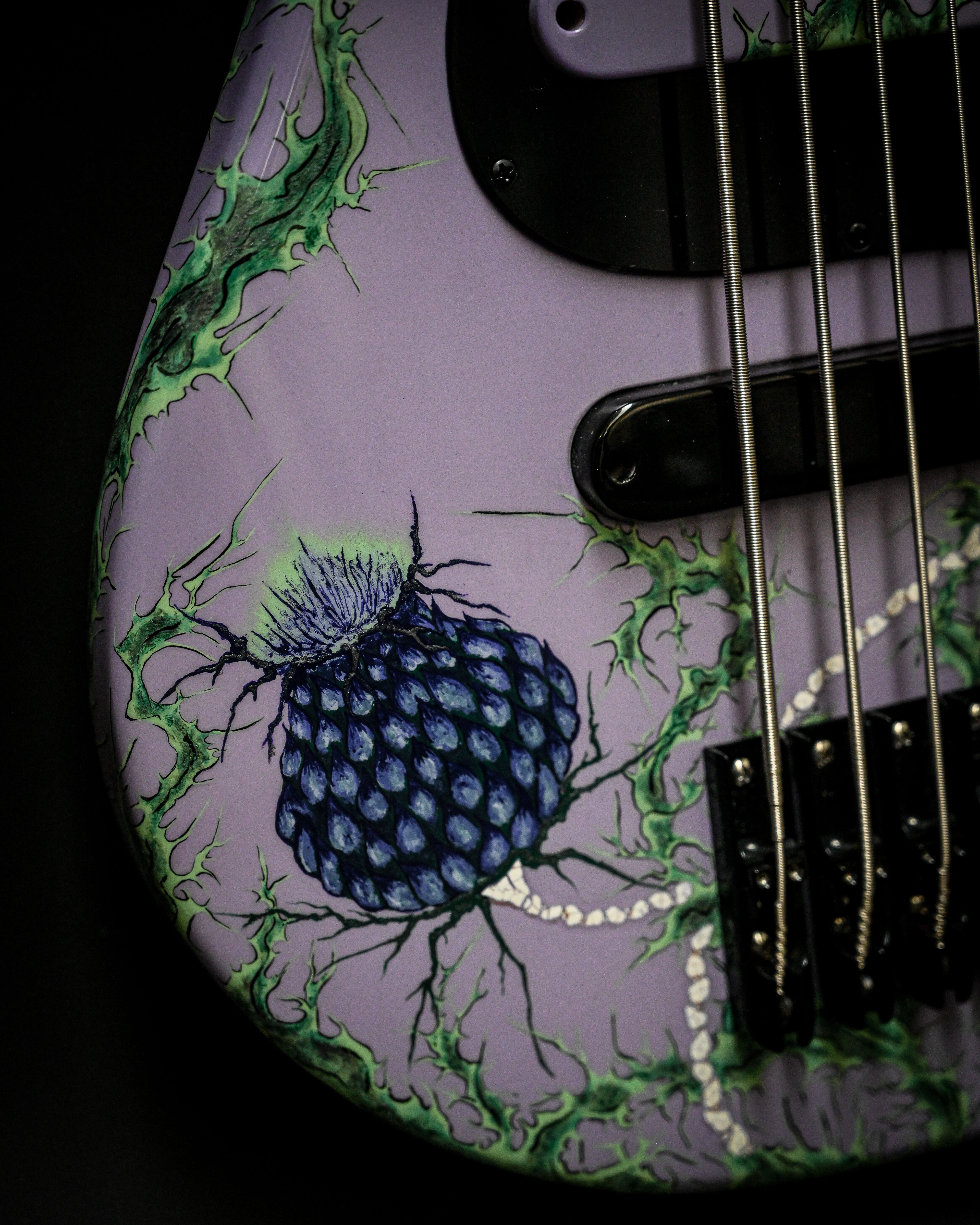 Gros plan sur le corps d'une guitare violette avec un motif de chardon bleu et des accents verts, avec des cordes en métal et des accessoires noirs.