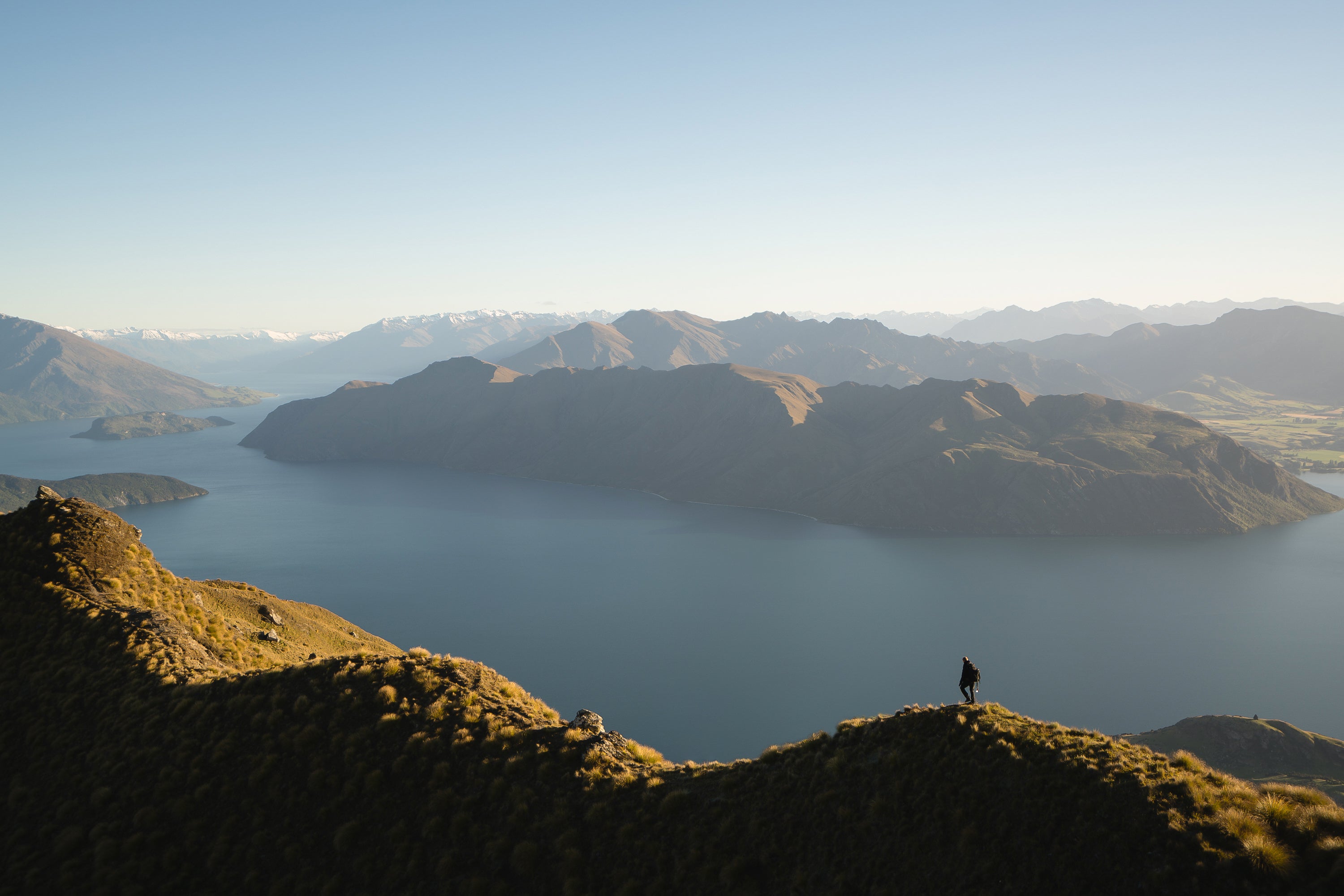 Un randonneur se tenant sur une crête surplombant un lac serein et des montagnes sous un ciel bleu clair
