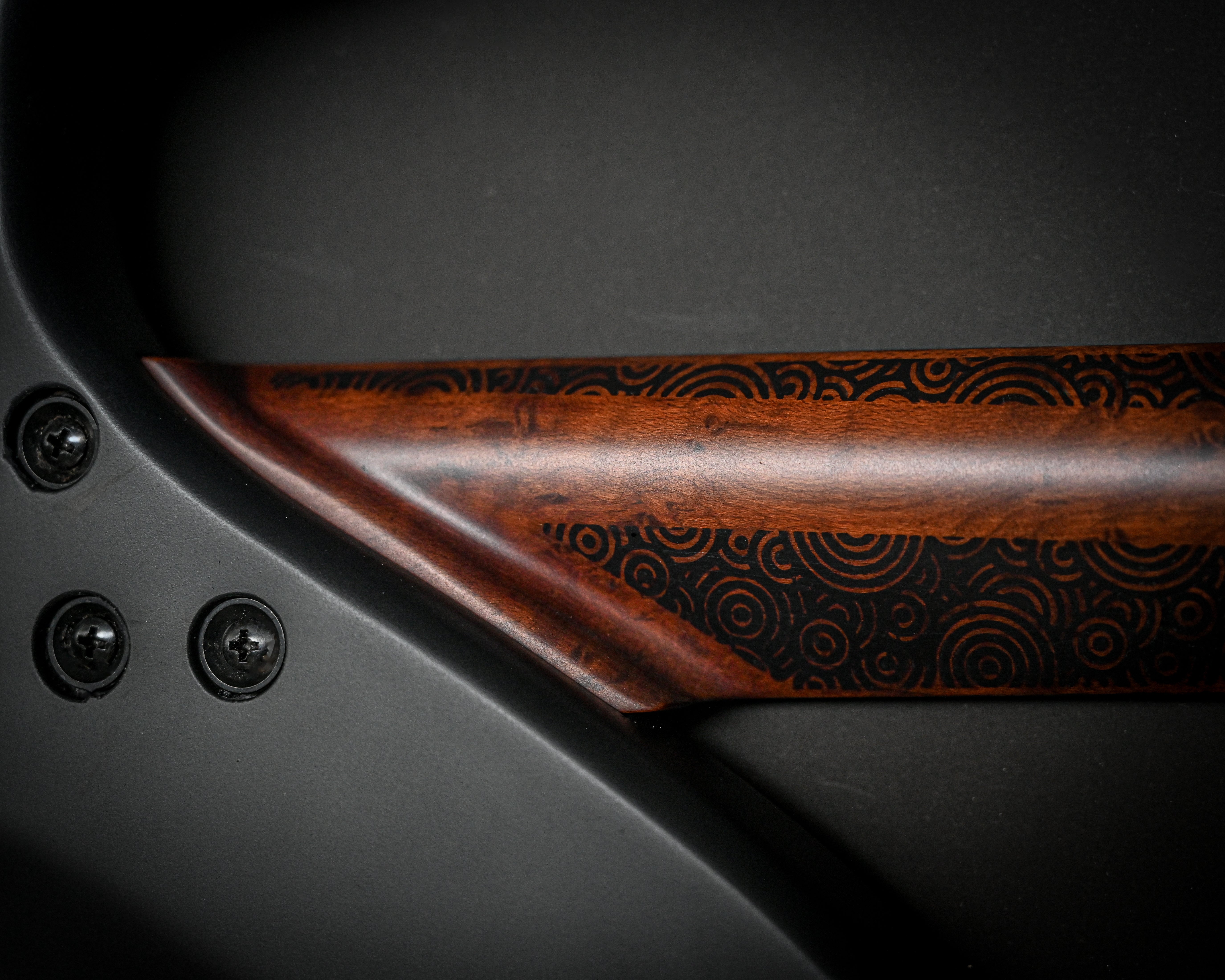 Close-up of a wooden guitar neck with intricate circular patterns engraved, contrasted against a black background and screws.