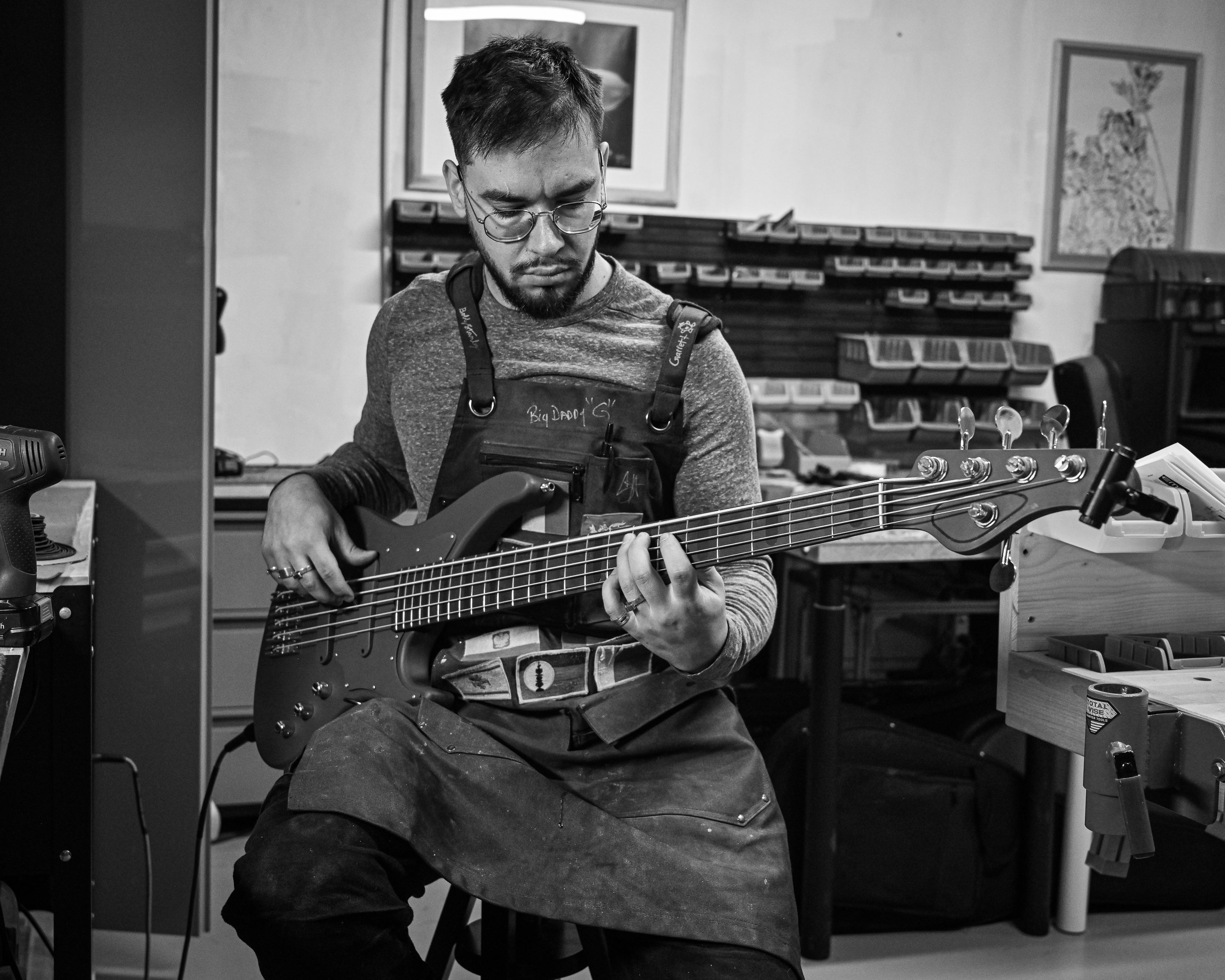 Luthier tocando y afinando un bajo en un taller, rodeado de herramientas y partes de guitarra, dedicado al trabajo artesanal.